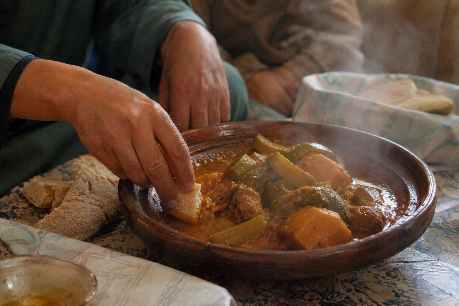 Hands breaking bread and dipping it into a shared Moroccan tajine