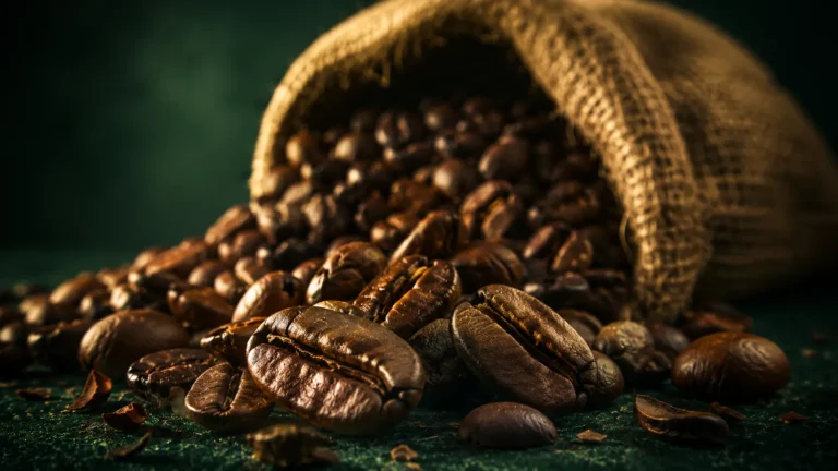 Roasted Yemeni coffee beans spilling from a burlap sack on a deep Meski-green surface.